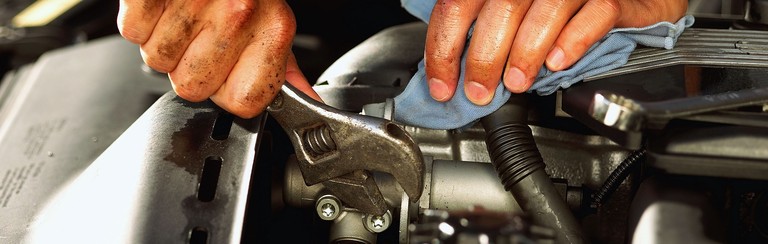Nahansicht der Hände eines Mechanikers mit einem Schraubenschlüssel über der offenen Motorhaube eines Autos.