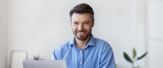 Ein junger Mann sitzt an einem Tisch, lächelt und arbeitet am Laptop. Im Hintergrund sind Pflanzen und ein Kaffeebecher zu sehen.