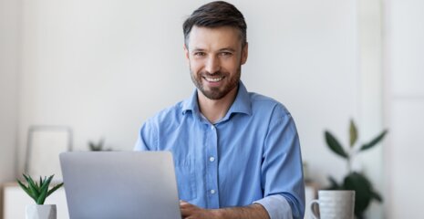 Ein junger Mann sitzt an einem Tisch, lächelt und arbeitet am Laptop. Im Hintergrund sind Pflanzen und ein Kaffeebecher zu sehen.