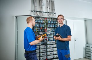 Zwei Techniker stehen in einem Elektroraum vor einem Schaltkasten. Einer hält ein Messgerät, der andere ein Tablet, beide arbeiten zusammen.