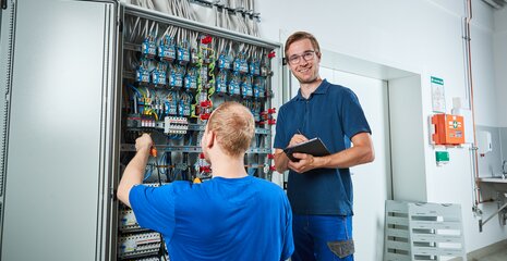 Zwei Techniker arbeiten an einem Schaltschrank. Einer löst Kabelverbindungen, während der andere Notizen macht. Beide wirken konzentriert.