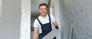 Ein Handwerker in blauer Latzhose und T-Shirt lächelt, während er eine Fliese in der Hand hält. Der Raum ist frisch renoviert.