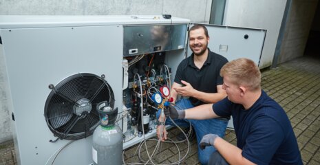 Zwei Techniker arbeiten an einer Klimaanlage. Einer erklärt und zeigt Messgeräte, während der andere an den Leitungen arbeitet.