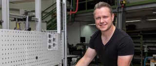 Ein junger Mann steht in einer Werkstatt und bearbeitet ein Metallstück. Er lächelt und trägt Arbeitshandschuhe. Werkzeug ist um ihn herum verteilt.
