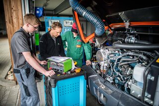 Drei Fachleute arbeiten an einem Motor in einer Werkstatt. Sie überprüfen technische Einrichtungen und diskutieren über die Reparatur.