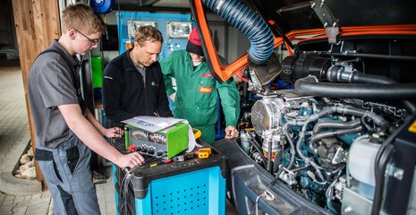 Drei Fachleute arbeiten an einem Motor in einer Werkstatt. Sie überprüfen technische Einrichtungen und diskutieren über die Reparatur.