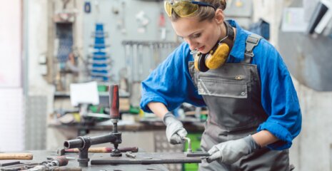 Eine junge Frau in Arbeitskleidung steht an einer Werkbank, konzentriert bei der Arbeit mit Werkzeugen. Sicherheitsbrille und Ohrenschutz tragen sie zum Schutz.