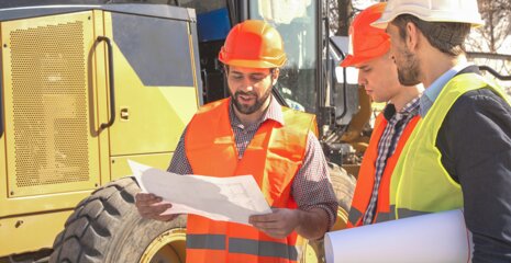 Drei Bauarbeiter in Sicherheitskleidung besprechen Pläne vor einer Baustelle. Im Hintergrund ist schweres Gerät zu sehen.