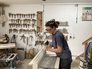 Eine Person arbeitet in einer Werkstatt, umgeben von Werkzeugen. Sie bearbeitet ein Holzstück mit viel Konzentration.