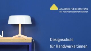 Das Bild zeigt eine moderne Tischlampe auf einem Holztisch vor einer blauen Wand. Daneben stehen Werkzeuge und Materialien für Handwerker:innen.